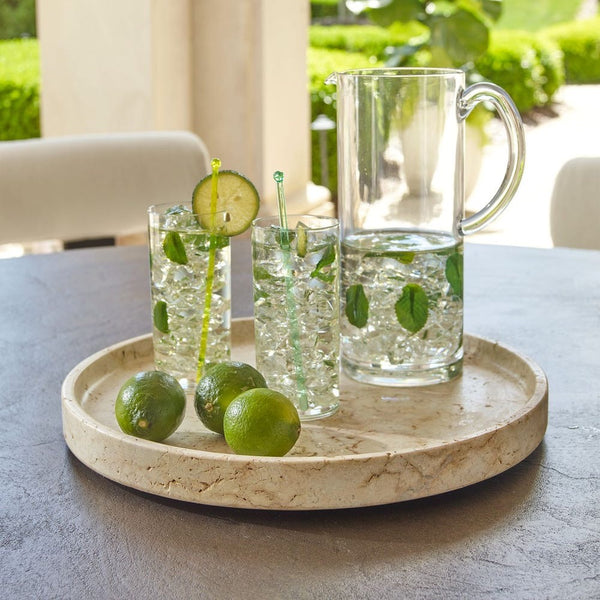 Clear glasses with lime slices and a pitcher on a stone tray outdoors.