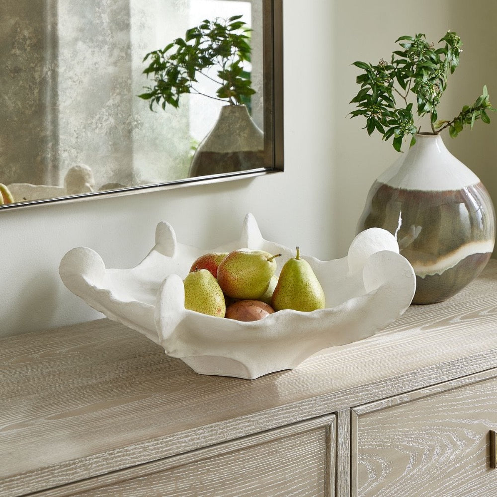 Decorative white bowl with pears on a wooden surface, next to a reflective mirror and a vase with greenery.
