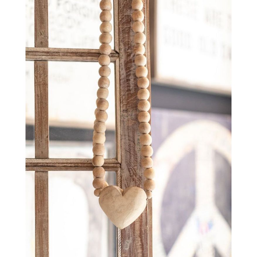 A string of Large Heart Wooden Prayer Beads by Sugarboo & Co., featuring an oversized heart-shaped pendant, adorns a rustic wooden window frame. In the blurred background, part of a peace sign is visible. The overall aesthetic is cozy and vintage—truly a unique home accessory.
