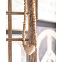 A string of Large Heart Wooden Prayer Beads by Sugarboo & Co., featuring an oversized heart-shaped pendant, adorns a rustic wooden window frame. In the blurred background, part of a peace sign is visible. The overall aesthetic is cozy and vintage—truly a unique home accessory.

