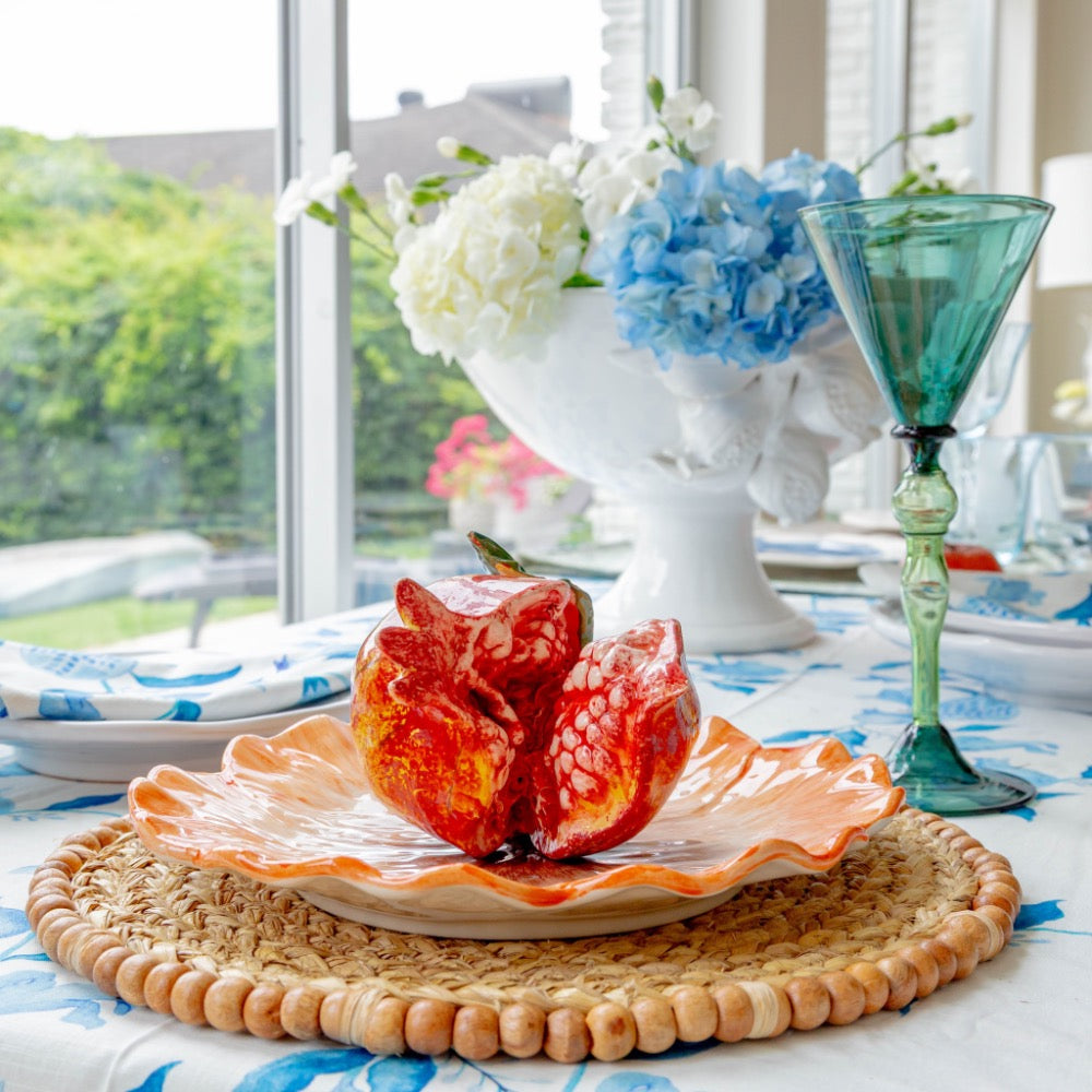 Decorative table setting with a pomegranate on a plate, flowers in the background, and a glass of water.