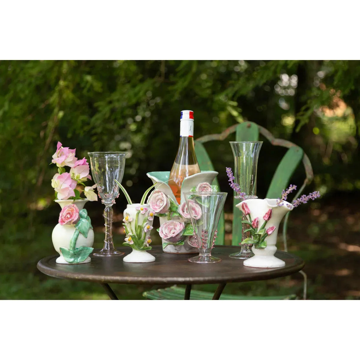 A round table is adorned with Abigails' Handmade White + Pink Rose Vase, glassware, and a wine bottle in the garden.
