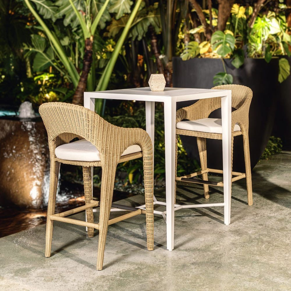 Two wicker bar stools with white cushions in front of a white table outdoors.