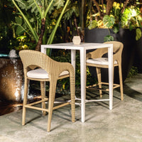 Two wicker bar stools with white cushions in front of a white table outdoors.