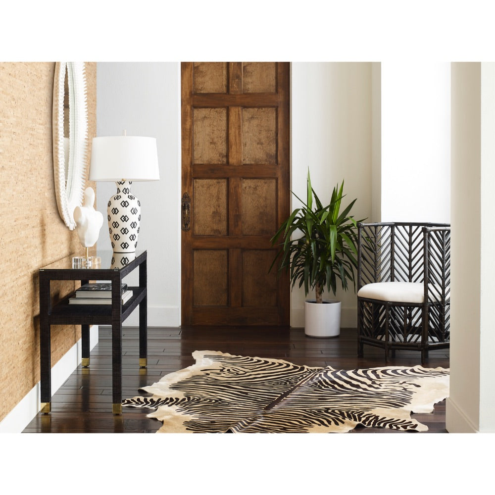 A contemporary entryway features a wooden door, zebra print rug, and a sleek black table adorned with a matte white lamp. The White Vogue Accent Sculpture by Wildwood Home introduces an artistic touch, complemented by a chair with a leaf pattern positioned beside the vibrant plant.