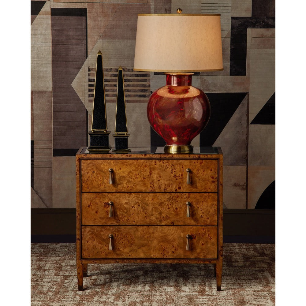 A three-drawer wooden nightstand with a rich burl wood grain finish and silver metal drawer pulls, styled in a room setting with a red glass table lamp, black decorative objects, and a geometric patterned wall.