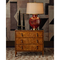 A three-drawer wooden nightstand with a rich burl wood grain finish and silver metal drawer pulls, styled in a room setting with a red glass table lamp, black decorative objects, and a geometric patterned wall.