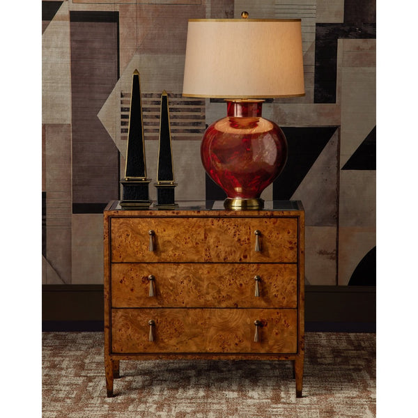 A three-drawer wooden nightstand with a rich burl wood grain finish and silver metal drawer pulls, styled in a room setting with a red glass table lamp, black decorative objects, and a geometric patterned wall.