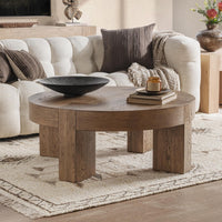 A round wooden coffee table with thick block legs in a warm medium brown finish, styled in a living room setting with a black bowl, books, and a candle on top, positioned in front of a beige tufted sofa.