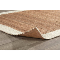 A close-up of a textured area rug with a warm brown center and cream-colored borders, shown partially folded over on a gray wood-look floor.