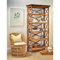 A tall rustic bamboo bookshelf with five shelves and crisscross side supports, styled in a room next to a woven rattan armchair with a cream pillow and a framed floral print on the wall.