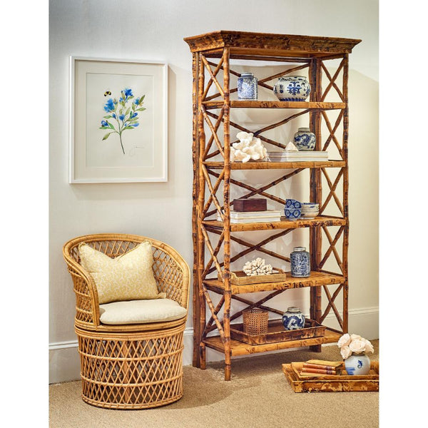 A tall rustic bamboo bookshelf with five shelves and crisscross side supports, styled in a room next to a woven rattan armchair with a cream pillow and a framed floral print on the wall.