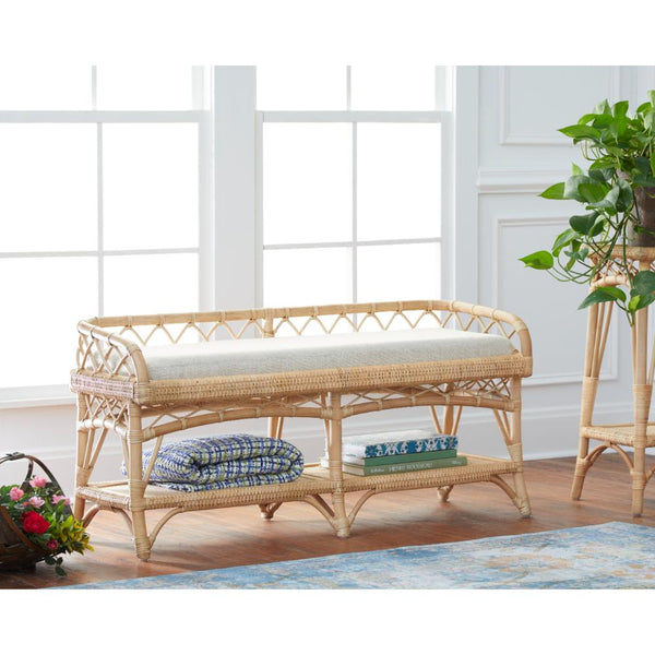 A natural rattan entryway bench with a light beige cushion, set in a bright room with wood floors, a blue patterned rug, and a potted plant on a side stand.