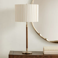 A table lamp with a cream pleated drum shade, a dark brown wooden stem, and brass-finished metal base and collar, shown on a dark wood console table against a light wall with a decorative gold mirror.