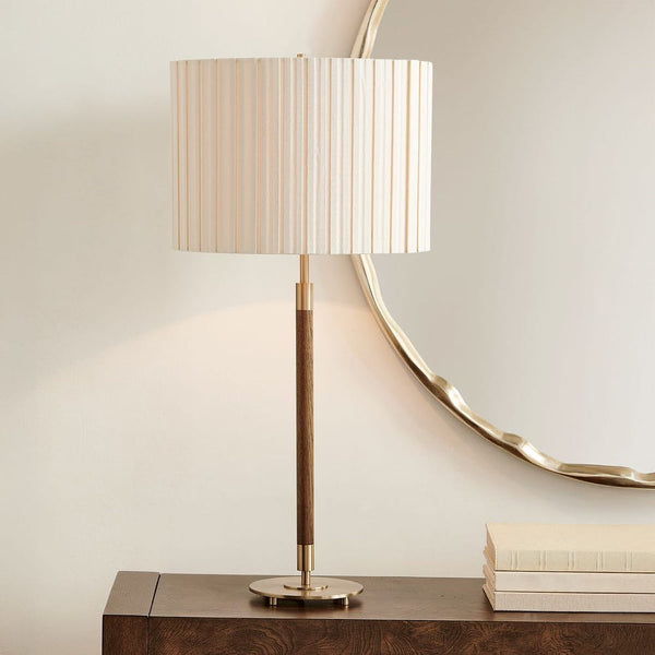 A table lamp with a cream pleated drum shade, a dark wood central stem, and a brushed gold base, styled on a dark wooden console table next to a stack of books and a decorative mirror.