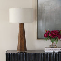 Decorative table lamp with a textured brown base on a dark surface, next to a vase of flowers and books.