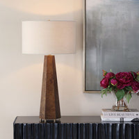 Decorative table lamp with a textured brown base and white shade on a dark surface, with a painting and flowers in the background.