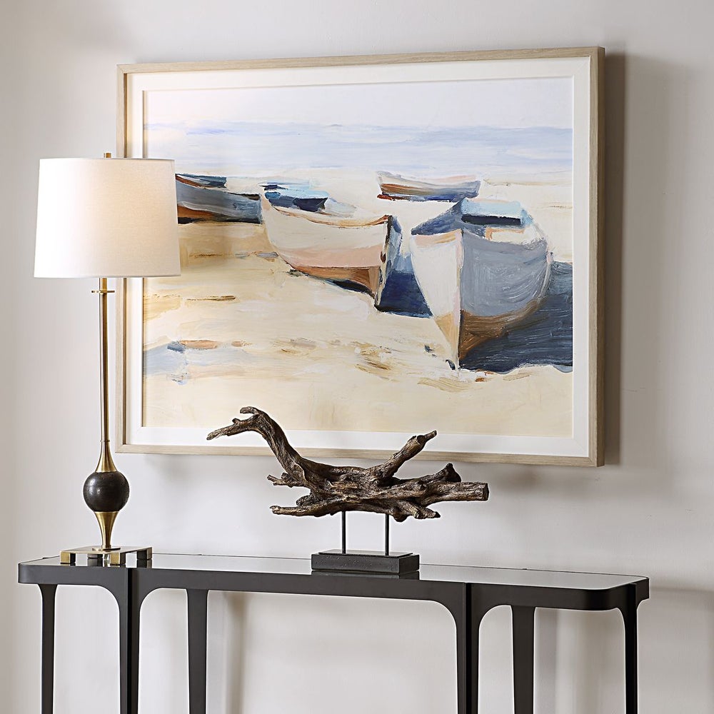 Table with lamp, driftwood sculpture, and Uttermost Beached Boats Framed Print create a tranquil atmosphere.