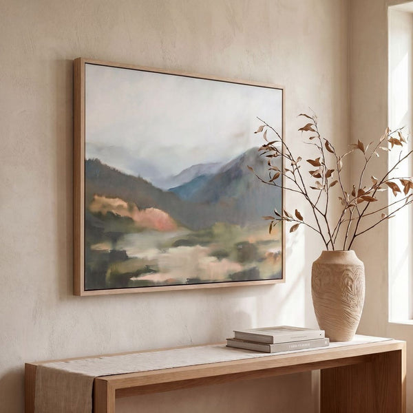Framed mountain landscape painting on a wall above a wooden console table with a vase and books.