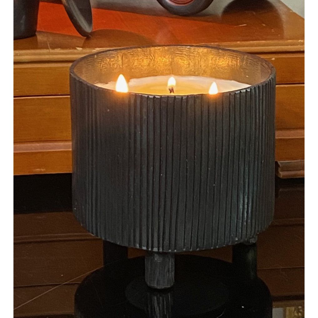 A lit Blacksmith Forged Candle by Himalayan Trading Post on a reflective surface, soy wax blend in ribbed black vessel.