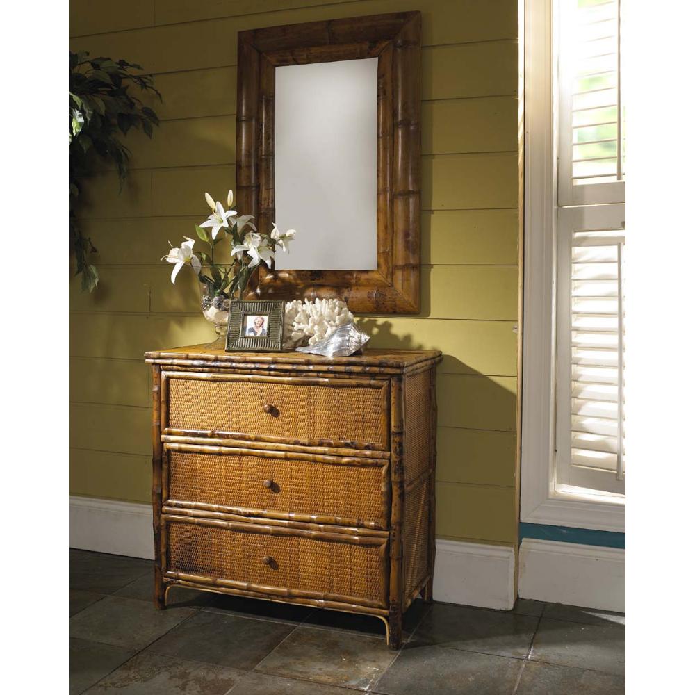 A three-drawer rattan chest of drawers with a bamboo-style frame, topped with a matching rectangular mirror, white lilies, and a small picture frame, set against a yellow-green paneled wall.