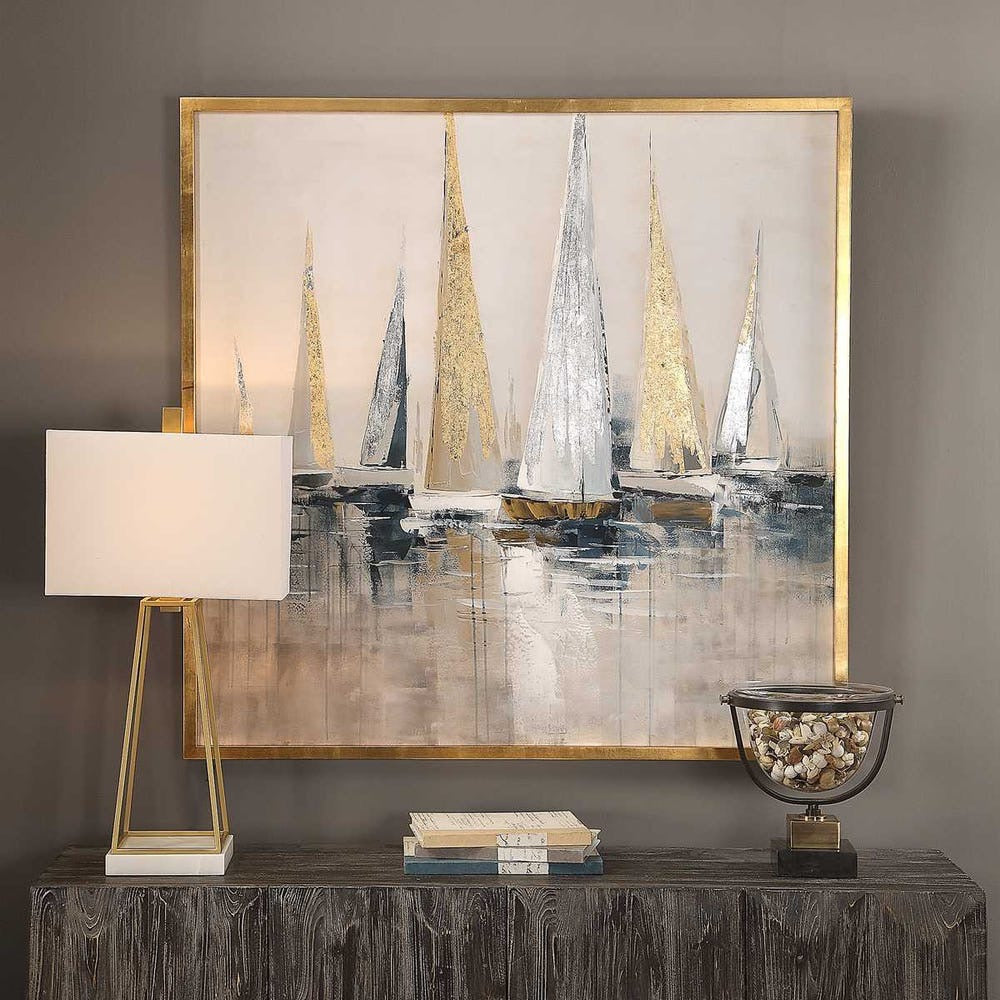 Abstract painting of sailboats with a gold frame, lamp, books, and decorative bowl on a wooden console table.