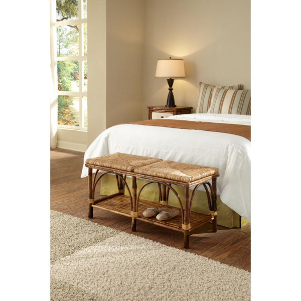 A woven rattan storage bench with a cushioned seat and lower shelf, styled in a bedroom next to a bed with white linens.