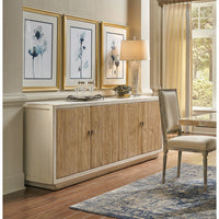 The elegant dining room showcases a Sarreid, Ltd. Antiqued Natural Pine Cabinet with a beige finish, adorned with a classic lamp and decorative items. Above the sideboard, three framed abstract paintings are displayed on the wall. A patterned rug and upholstered chair are also visible, while large windows with light-colored curtains allow natural light to fill the farmhouse-style room.