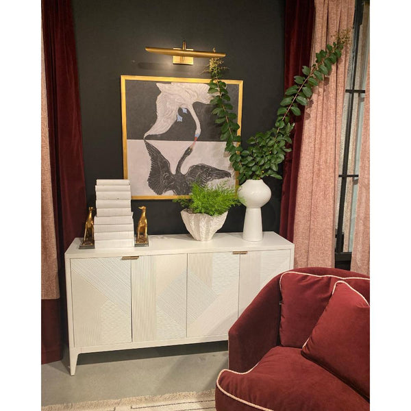 A white cabinet against a dark wall is adorned with "The Swan Framed Art" by Shadow Catchers Art and stacked books, while vases enhance its elegance. A red sofa in the foreground complements the bold design, creating a striking contrast that's both inviting and sophisticated.