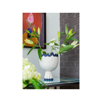 Decorative vase with green leaves and white flowers on a glass table, with a colorful abstract painting in the background.