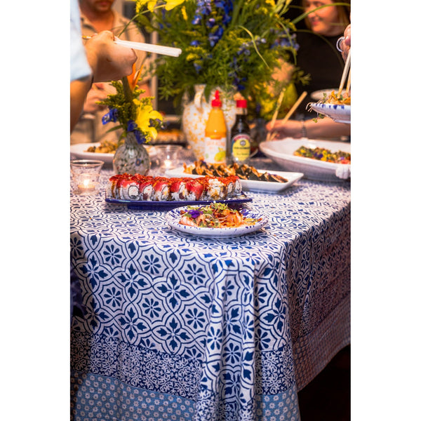 A table with sushi, flowers, and condiments on an Abigails Beach Chic Cabana Tablecloth, surrounded by guests serving themselves.