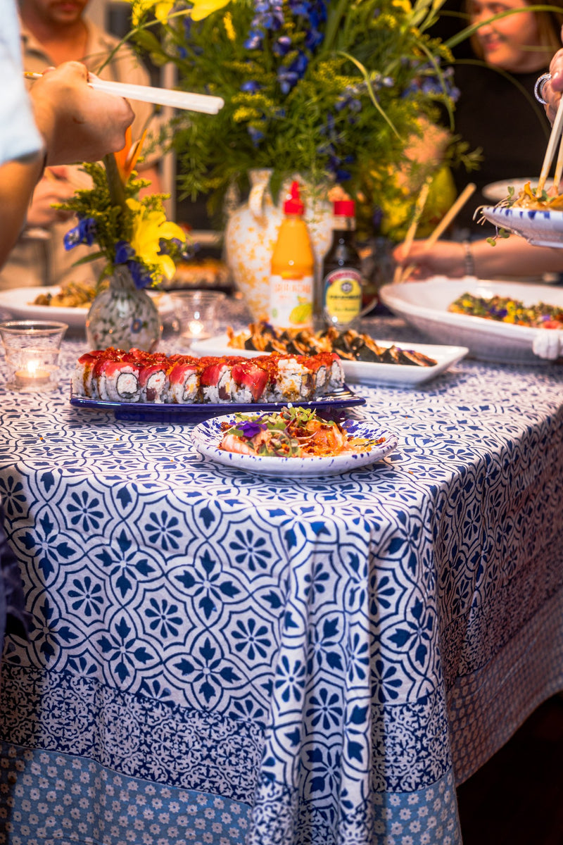 A table with sushi, flowers, and condiments on an Abigails Beach Chic Cabana Tablecloth, surrounded by guests serving themselves.