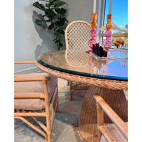A round dining table with a woven rattan base and clear glass top, surrounded by matching woven chairs in a bright room with a window and pink glass candlesticks on the table.