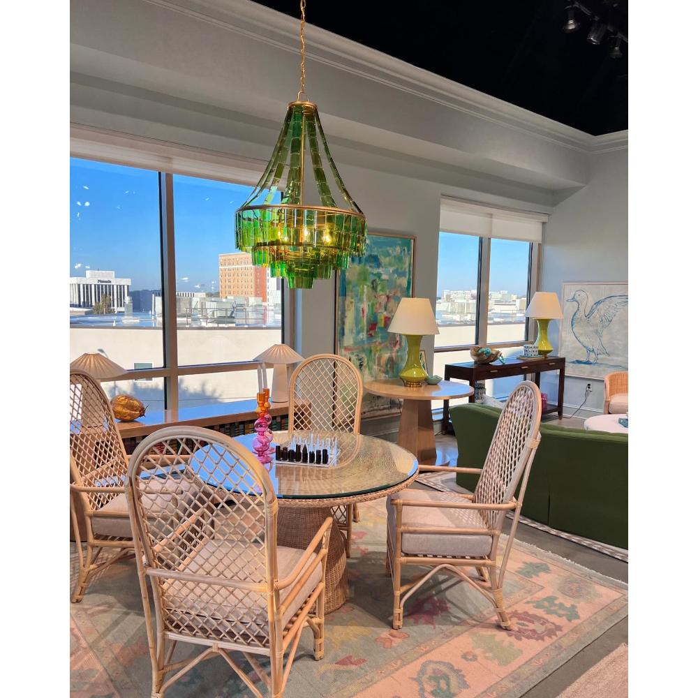 A green glass chandelier hangs over a round glass-top dining table with four woven rattan chairs in a bright, modern dining room with large windows and city views.