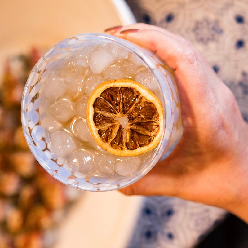 Hand holding an Abigails Torcello Spotted White Rosa Tumbler with ice and a dried lemon slice over a patterned surface.