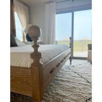 A wooden bed frame with a turned spindle head and footboard, dressed in a textured cream quilt, shown in a bedroom setting with a shag rug and large windows looking out to the beach.