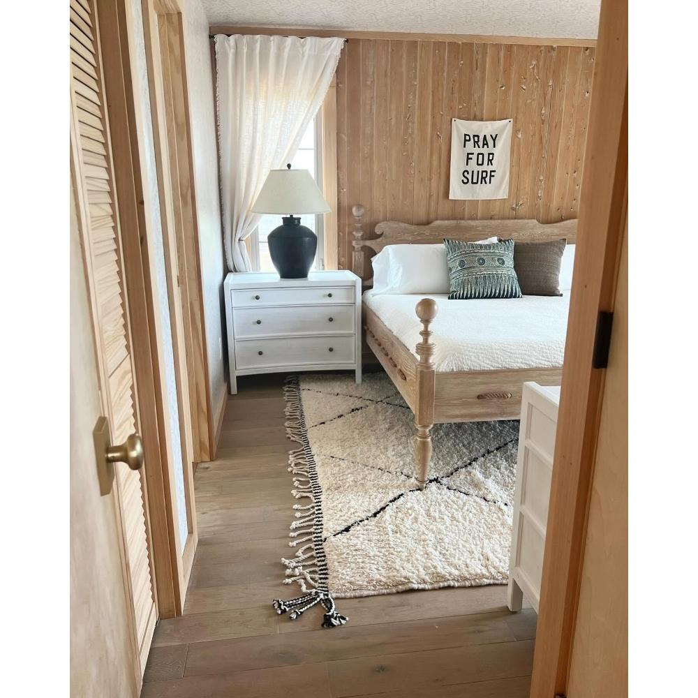 A rustic bedroom interior with a light wood bed, white bedding, a white nightstand, a black table lamp, a cream rug, and wood-paneled walls. A small wall sign reads “PRAY FOR SURF.