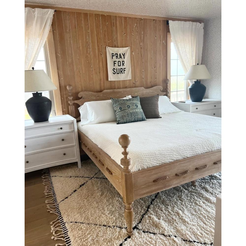 A rustic light wood four-poster bed with white bedding, styled in a bedroom with a wood-paneled wall, white nightstands, black table lamps, and a cream shag rug.