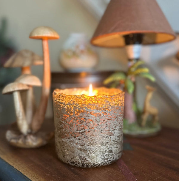 A lit candle in a textured, metallic silver and gold crimped glass holder sits on a wooden table, surrounded by decorative items including wooden mushrooms, a lamp, and a small figurine in a warm, cozy indoor setting.