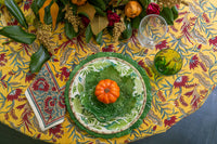 Decorative table setting with a green plate, small pumpkin, and glass on a yellow floral tablecloth.