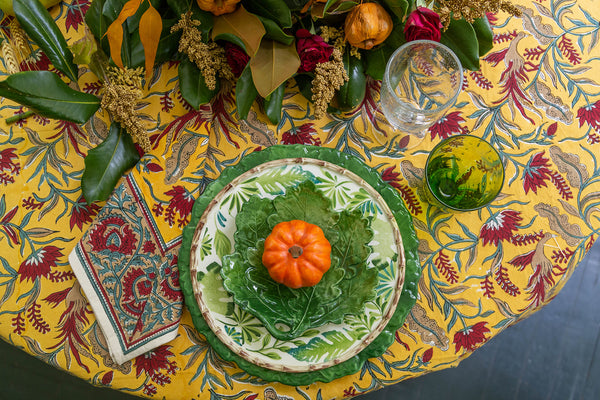 Decorative table setting with a green plate, small pumpkin, and glass on a yellow floral tablecloth.