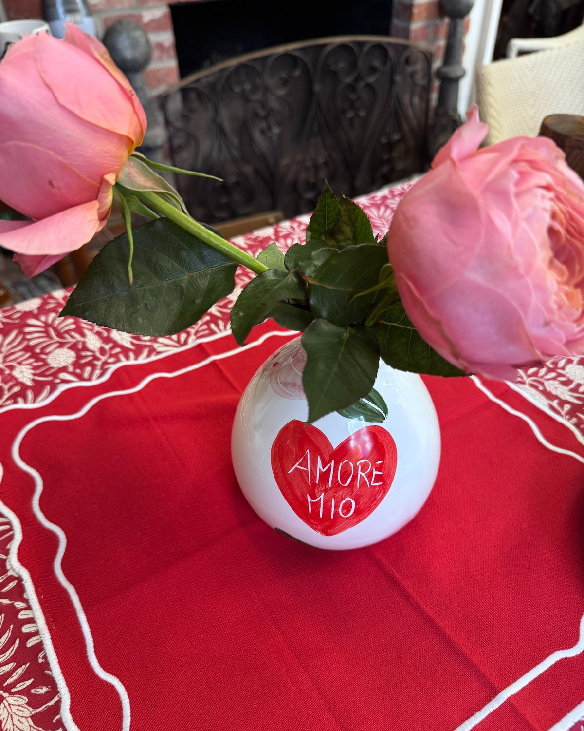 White vase with red heart and 'AMORE MIO' text on a red tablecloth with pink roses.