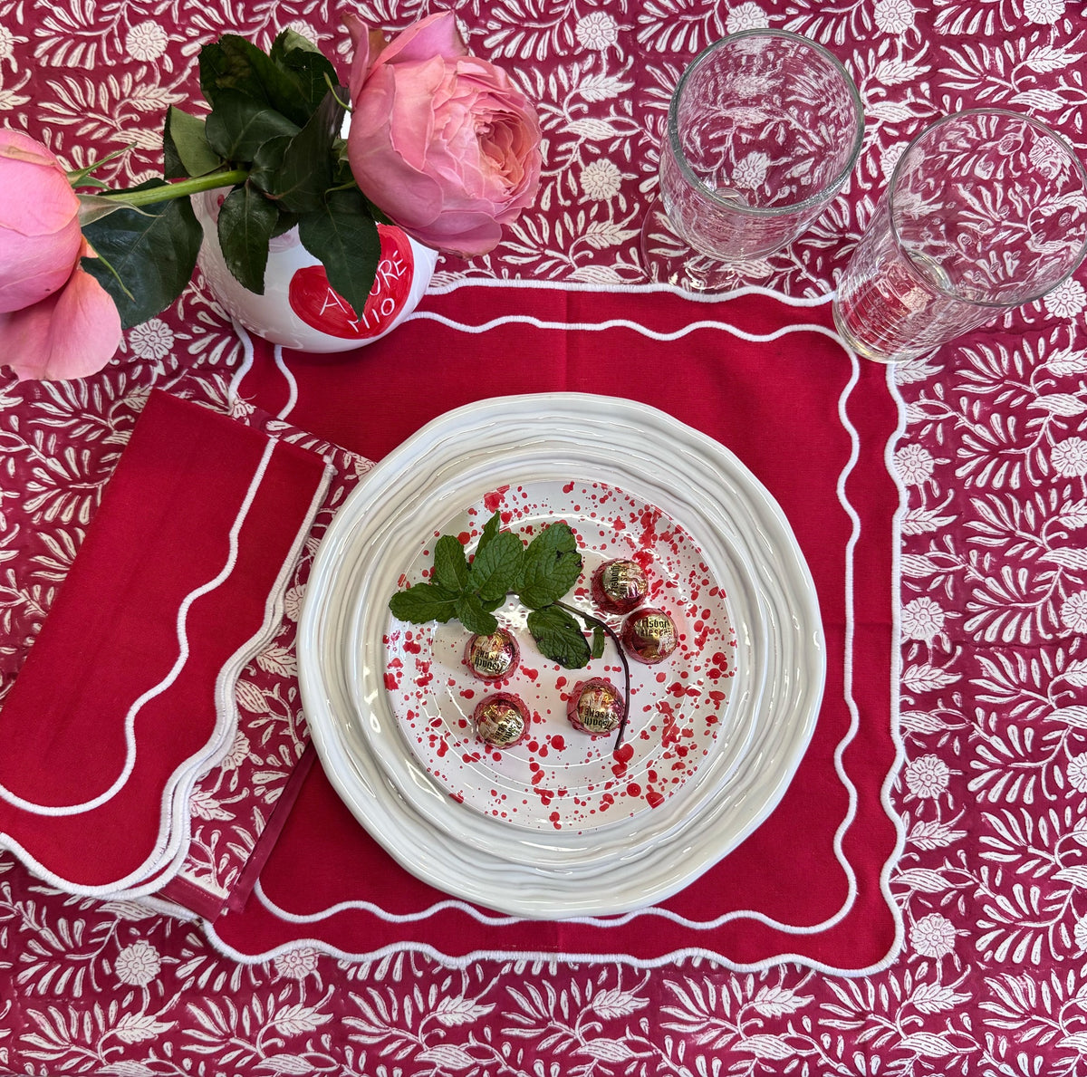 Table setting with a plate of food, glasses, and a pink flower on a red patterned tablecloth.