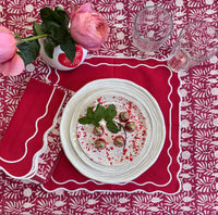 Table setting with a plate of food, glasses, and a pink flower on a red patterned tablecloth.