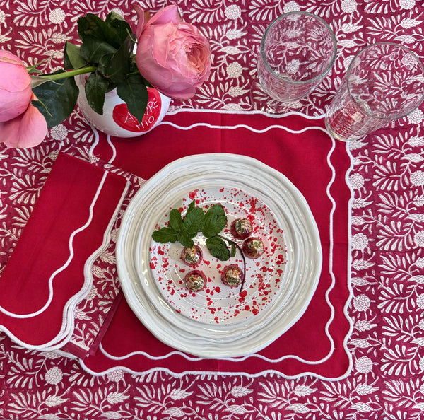 Table setting with a plate of food, glasses, and a pink flower on a red patterned tablecloth.