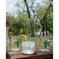 Clear pitcher and glasses with cactus designs on a wooden surface outdoors.