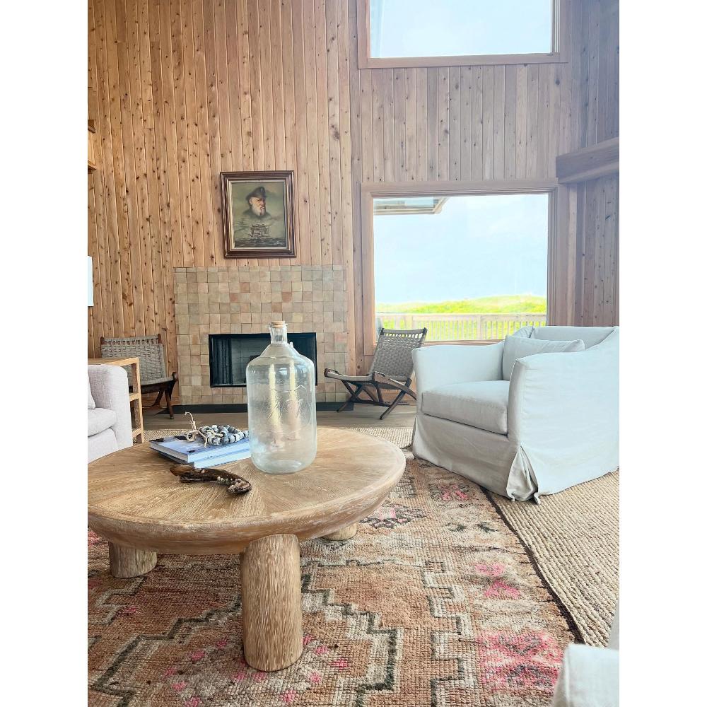 A rustic round wooden coffee table in a coastal-style living room with wood-paneled walls, a fireplace, a light gray armchair, and a large clear glass bottle on top.