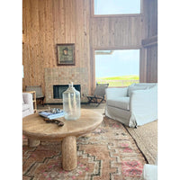 A rustic round wooden coffee table in a coastal-style living room with wood-paneled walls, a fireplace, a light gray armchair, and a large clear glass bottle on top.