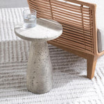 A glass of water sits on the Collins Concrete Accent Table by Classic Home next to rust outdoor furniture and a rug.