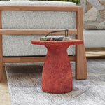 A Collins Concrete Accent Table by Classic Home (rust color) holds books and glasses beside a wood-framed gray sofa.